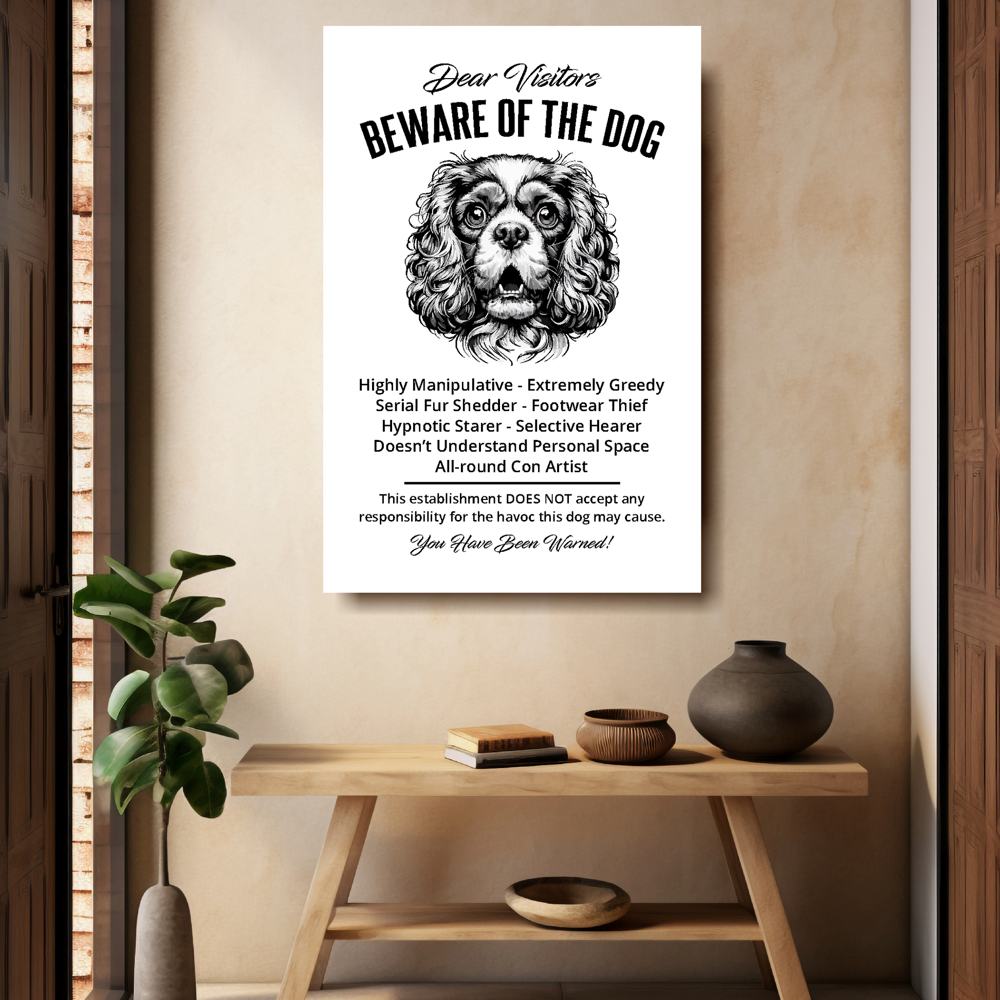 A funny Cavalier King Charles Spaniel 'Beware of the Dog' disclaimer notice. Black text and dog image on a white background. Mounted on a beige wall above a wooden bench.